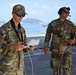 Two U.S. Coast Guard personnel in OCP (Operational Camouflage Pattern) uniforms are operating equipment at what appears to be a port or waterfront facility. The individual in the foreground, identified by name tape as 'KLAUS,' is handling a white device with pink cables, while 'LOCKWOOD' in the background wears a communications headset. Both are wearing sunglasses and their uniforms display U.S. Coast Guard branch tape and American flag patches.