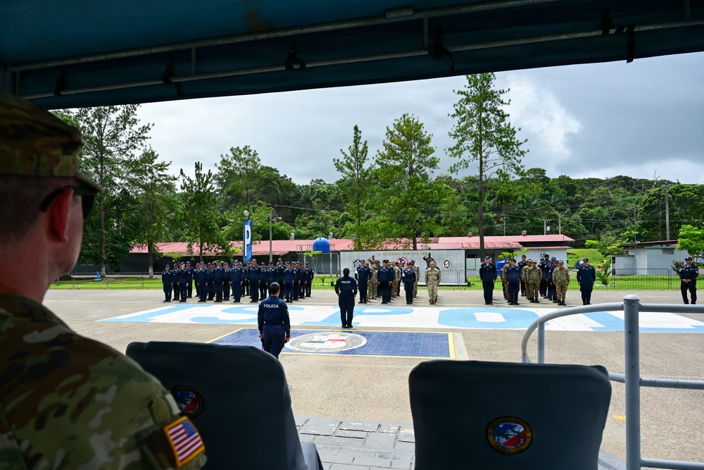 A military or law enforcement ceremony is taking place on an outdoor parade ground, viewed from a covered observation area. Personnel in blue police uniforms ('POLICIA') and military camouflage uniforms are assembled in formation, with a large painted emblem visible on the ground. A U.S. Army soldier wearing an American flag patch is visible in the foreground observing the event.