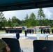 A military or law enforcement ceremony is taking place on an outdoor parade ground, viewed from a covered observation area. Personnel in blue police uniforms ('POLICIA') and military camouflage uniforms are assembled in formation, with a large painted emblem visible on the ground. A U.S. Army soldier wearing an American flag patch is visible in the foreground observing the event.