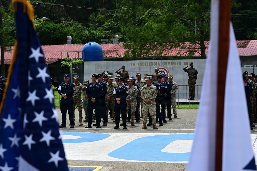 A formal military ceremony or parade is taking place at what appears to be a Latin American military installation, with personnel in mixed uniforms standing in formation. The Spanish inscription 'La Excelencia Comienza Aquí' (Excellence Begins Here) is visible on a wall in the background, along with bronze statues of saluting soldiers. A US flag is visible in the foreground alongside what appears to be a blue-and-white flag, suggesting a joint or bilateral military event.