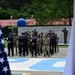 A formal military ceremony or parade is taking place at what appears to be a Latin American military installation, with personnel in mixed uniforms standing in formation. The Spanish inscription 'La Excelencia Comienza Aquí' (Excellence Begins Here) is visible on a wall in the background, along with bronze statues of saluting soldiers. A US flag is visible in the foreground alongside what appears to be a blue-and-white flag, suggesting a joint or bilateral military event.