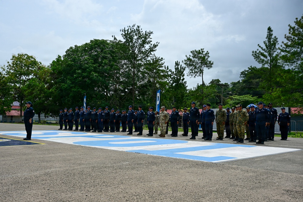 A formal military or law enforcement parade formation is taking place on an outdoor paved area featuring large blue and white painted letters or markings on the ground. The majority of personnel are dressed in dark navy blue uniforms with caps, while a smaller group in the center-right wears camouflage military attire. The event appears to be an official ceremony, possibly a graduation, inspection, or joint exercise involving multiple security forces.