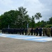 A formal military or law enforcement parade formation is taking place on an outdoor paved area featuring large blue and white painted letters or markings on the ground. The majority of personnel are dressed in dark navy blue uniforms with caps, while a smaller group in the center-right wears camouflage military attire. The event appears to be an official ceremony, possibly a graduation, inspection, or joint exercise involving multiple security forces.