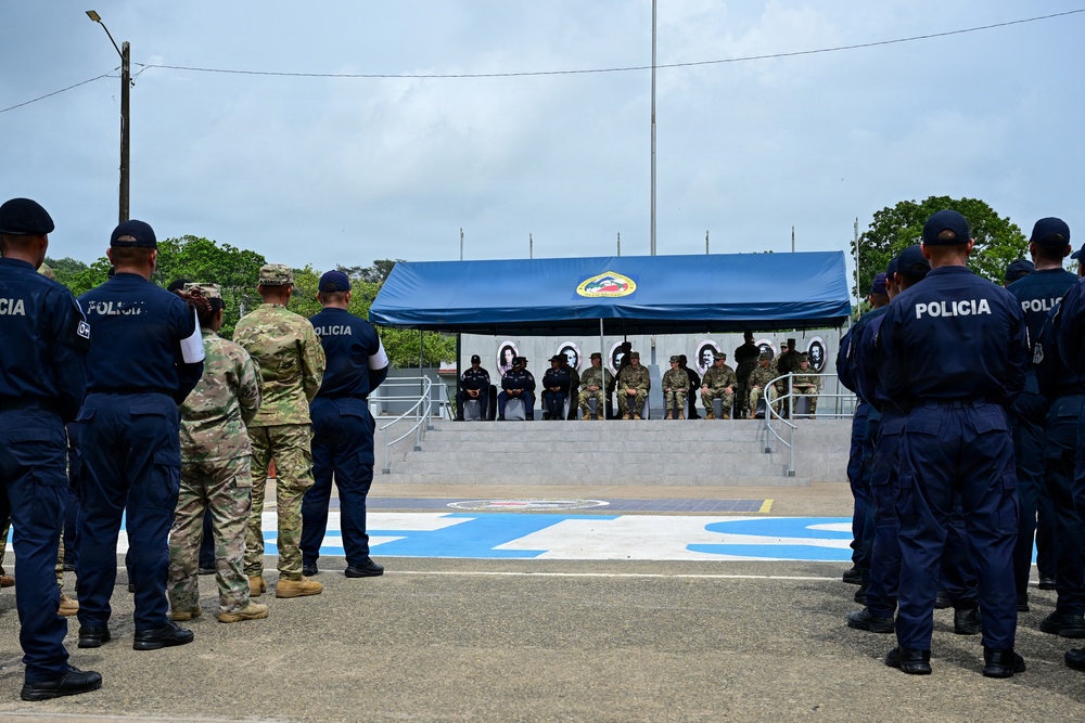 A formal military and police ceremony is taking place outdoors at what appears to be a Latin American police facility. Personnel wearing 'POLICIA' uniforms in dark blue stand in formation facing a covered reviewing stand where seated dignitaries, including both camouflage-uniformed military personnel and civilian/police officials, are presiding. The setting includes a blue canopy with an emblem, portrait photographs displayed in the background, and painted markings on the ground.