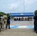 A formal military and police ceremony is taking place outdoors at what appears to be a Latin American police facility. Personnel wearing 'POLICIA' uniforms in dark blue stand in formation facing a covered reviewing stand where seated dignitaries, including both camouflage-uniformed military personnel and civilian/police officials, are presiding. The setting includes a blue canopy with an emblem, portrait photographs displayed in the background, and painted markings on the ground.