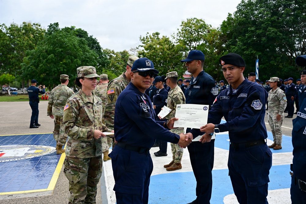 A military/police graduation ceremony is taking place outdoors on a decorated parade ground. A senior officer in dark blue uniform is presenting a 'Certificado de Finalización' (Certificate of Completion) to a recipient, while U.S. Army personnel in MultiCam uniforms observe. Multiple uniformed personnel from what appears to be a Latin American national police or military force are standing in formation in the background.