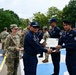A military/police graduation ceremony is taking place outdoors on a decorated parade ground. A senior officer in dark blue uniform is presenting a 'Certificado de Finalización' (Certificate of Completion) to a recipient, while U.S. Army personnel in MultiCam uniforms observe. Multiple uniformed personnel from what appears to be a Latin American national police or military force are standing in formation in the background.