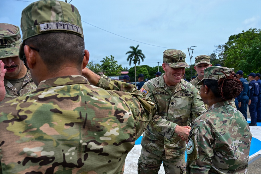 A group of U.S. Army soldiers in OCP (Operational Camouflage Pattern) uniforms are engaged in what appears to be a military ceremony or greeting event outdoors. A smiling officer identified by nametape as 'CLEMENS' with a Lieutenant Colonel oak leaf insignia is shaking hands with a female soldier, while another soldier identified as 'J. PITTY' faces away from the camera. The setting appears to be in a tropical location, possibly Central America or the Caribbean, with local police or security forces visible in the background.