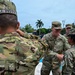 A group of U.S. Army soldiers in OCP (Operational Camouflage Pattern) uniforms are engaged in what appears to be a military ceremony or greeting event outdoors. A smiling officer identified by nametape as 'CLEMENS' with a Lieutenant Colonel oak leaf insignia is shaking hands with a female soldier, while another soldier identified as 'J. PITTY' faces away from the camera. The setting appears to be in a tropical location, possibly Central America or the Caribbean, with local police or security forces visible in the background.
