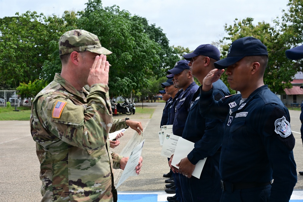 A U.S. Army soldier in OCP camouflage uniform is exchanging salutes with a line of personnel wearing dark navy blue uniforms and caps, who are holding certificates. The ceremony appears to be a graduation or recognition event, likely a joint training exercise between U.S. military and a Latin American law enforcement or security force. The event is taking place outdoors on a paved area with trees and buildings visible in the background.