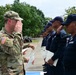 A U.S. Army soldier in OCP camouflage uniform is exchanging salutes with a line of personnel wearing dark navy blue uniforms and caps, who are holding certificates. The ceremony appears to be a graduation or recognition event, likely a joint training exercise between U.S. military and a Latin American law enforcement or security force. The event is taking place outdoors on a paved area with trees and buildings visible in the background.