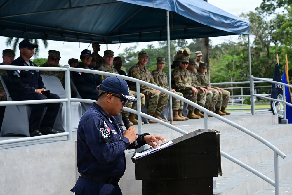 A uniformed official, wearing a navy blue uniform with a patch reading 'DICCOP' and a Panamanian flag insignia, is speaking at a podium during what appears to be a formal military or law enforcement ceremony. Behind him on a raised platform under a blue canopy, two groups are seated: personnel in dark uniforms on the left and soldiers in camouflage (OCP/MultiCam pattern) on the right, suggesting a joint multinational event. A flag and a sign reading 'PCL' are visible in the background.
