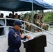 A uniformed official, wearing a navy blue uniform with a patch reading 'DICCOP' and a Panamanian flag insignia, is speaking at a podium during what appears to be a formal military or law enforcement ceremony. Behind him on a raised platform under a blue canopy, two groups are seated: personnel in dark uniforms on the left and soldiers in camouflage (OCP/MultiCam pattern) on the right, suggesting a joint multinational event. A flag and a sign reading 'PCL' are visible in the background.