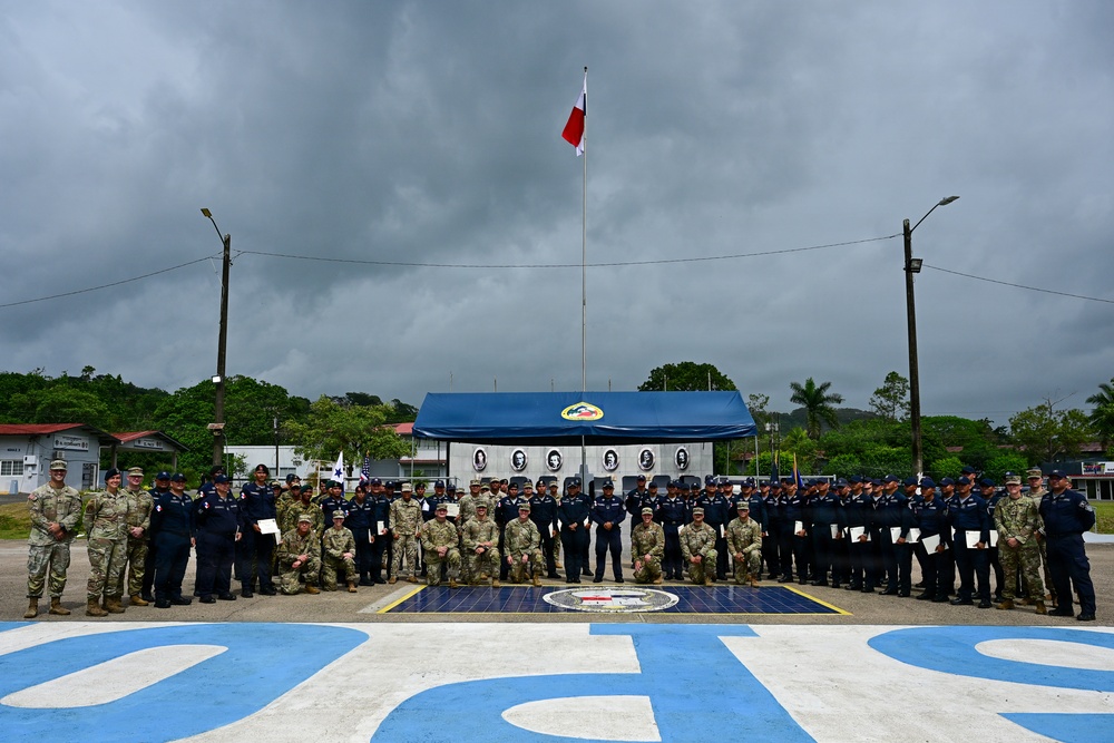 A large group of military and law enforcement personnel from multiple nations pose for a formal group photograph at what appears to be a joint training or ceremonial event. The setting includes a monument or memorial structure with portrait reliefs, a flagpole flying a red and white flag, and large blue lettering on the ground. Personnel are wearing a mix of camouflage military uniforms and dark blue uniforms, suggesting a multinational cooperative exercise or graduation ceremony.