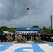 A large group of military and law enforcement personnel from multiple nations pose for a formal group photograph at what appears to be a joint training or ceremonial event. The setting includes a monument or memorial structure with portrait reliefs, a flagpole flying a red and white flag, and large blue lettering on the ground. Personnel are wearing a mix of camouflage military uniforms and dark blue uniforms, suggesting a multinational cooperative exercise or graduation ceremony.