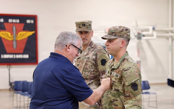 A civilian man in a blue polo shirt is pinning something onto the uniform of a U.S. Army soldier in OCP (Operational Camouflage Pattern) uniform, likely during a promotion or award ceremony. A second soldier wearing captain rank insignia (two silver bars) stands in the background observing. The event appears to take place inside a military armory or drill hall.