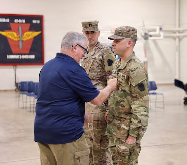 A civilian man in a blue polo shirt is pinning something onto the uniform of a U.S. Army soldier in OCP (Operational Camouflage Pattern) uniform, likely during a promotion or award ceremony. A second soldier wearing captain rank insignia (two silver bars) stands in the background observing. The event appears to take place inside a military armory or drill hall.