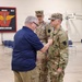 A civilian man in a blue polo shirt is pinning something onto the uniform of a U.S. Army soldier in OCP (Operational Camouflage Pattern) uniform, likely during a promotion or award ceremony. A second soldier wearing captain rank insignia (two silver bars) stands in the background observing. The event appears to take place inside a military armory or drill hall.