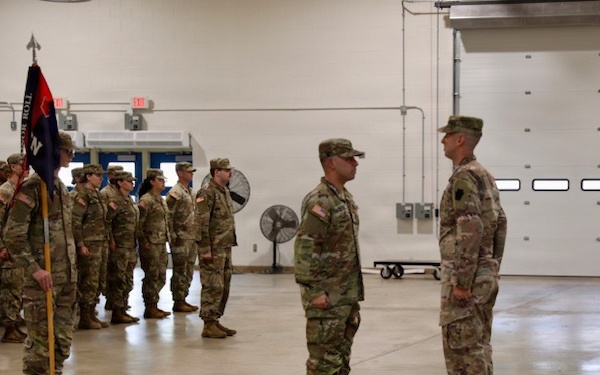 A military formation ceremony is taking place inside a large indoor facility, likely an armory or motor pool bay. Two soldiers stand facing each other in the foreground, possibly a commander and a reviewing officer, while a formation of soldiers stands at attention in the background with a unit guidon. The setting and uniforms suggest a U.S. Army National Guard or Reserve unit ceremony.