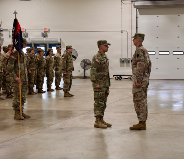 A military formation ceremony is taking place inside a large indoor facility, likely an armory or motor pool bay. Two soldiers stand facing each other in the foreground, possibly a commander and a reviewing officer, while a formation of soldiers stands at attention in the background with a unit guidon. The setting and uniforms suggest a U.S. Army National Guard or Reserve unit ceremony.