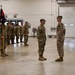 A military formation ceremony is taking place inside a large indoor facility, likely an armory or motor pool bay. Two soldiers stand facing each other in the foreground, possibly a commander and a reviewing officer, while a formation of soldiers stands at attention in the background with a unit guidon. The setting and uniforms suggest a U.S. Army National Guard or Reserve unit ceremony.