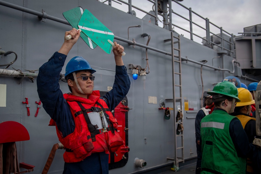 A naval crew member wearing a blue hard hat, sunglasses, and a red life vest is holding up two green paddle-shaped signaling boards above their head on the deck of a gray military vessel. Several other crew members in the background are wearing various colored hard hats (green, yellow, blue) and safety vests. The scene appears to depict a replenishment-at-sea (RAS) or underway replenishment operation.