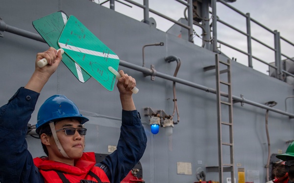 A naval crew member wearing a blue hard hat, sunglasses, and a red life vest is holding up two green paddle-shaped signaling boards above their head on the deck of a gray military vessel. Several other crew members in the background are wearing various colored hard hats (green, yellow, blue) and safety vests. The scene appears to depict a replenishment-at-sea (RAS) or underway replenishment operation.