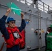 A naval crew member wearing a blue hard hat, sunglasses, and a red life vest is holding up two green paddle-shaped signaling boards above their head on the deck of a gray military vessel. Several other crew members in the background are wearing various colored hard hats (green, yellow, blue) and safety vests. The scene appears to depict a replenishment-at-sea (RAS) or underway replenishment operation.