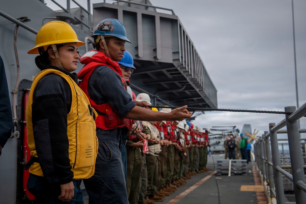 A line of naval personnel standing on the deck of a military vessel, holding a rope or line in a coordinated formation. The crew members are wearing safety equipment including hard hats in yellow and blue colors, and red or yellow life vests. This appears to be a standard naval deck operation, likely a mooring, replenishment at sea, or line-handling evolution.