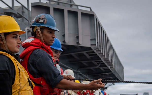 A line of naval personnel standing on the deck of a military vessel, holding a rope or line in a coordinated formation. The crew members are wearing safety equipment including hard hats in yellow and blue colors, and red or yellow life vests. This appears to be a standard naval deck operation, likely a mooring, replenishment at sea, or line-handling evolution.