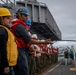 A line of naval personnel standing on the deck of a military vessel, holding a rope or line in a coordinated formation. The crew members are wearing safety equipment including hard hats in yellow and blue colors, and red or yellow life vests. This appears to be a standard naval deck operation, likely a mooring, replenishment at sea, or line-handling evolution.