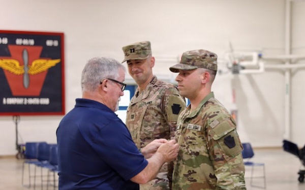 A civilian man in a blue polo shirt is pinning a medal or insignia onto a U.S. Army soldier in OCP (Operational Camouflage Pattern) uniform, while a second uniformed soldier stands behind observing. The ceremony appears to be taking place inside a military armory or hangar facility. A unit insignia banner is visible on the wall in the background, consistent with an Army aviation unit.