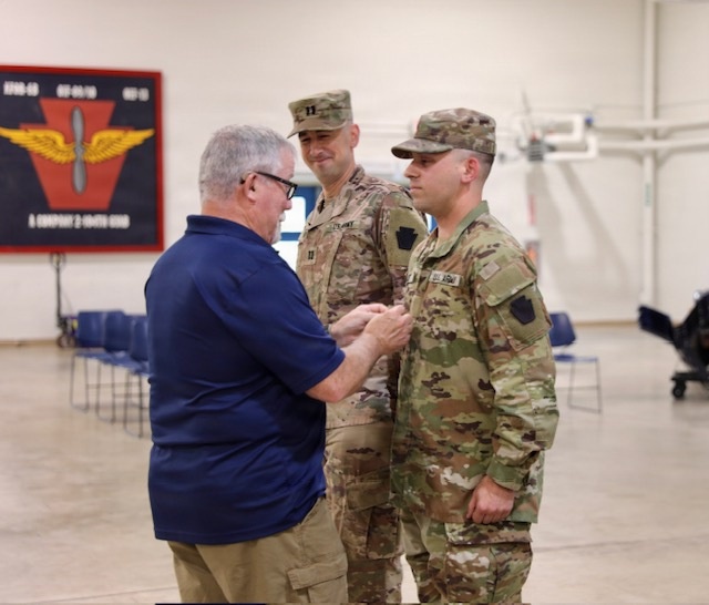 A civilian man in a blue polo shirt is pinning a medal or insignia onto a U.S. Army soldier in OCP (Operational Camouflage Pattern) uniform, while a second uniformed soldier stands behind observing. The ceremony appears to be taking place inside a military armory or hangar facility. A unit insignia banner is visible on the wall in the background, indicating an aviation unit.