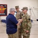A civilian man in a blue polo shirt is pinning a medal or insignia onto a U.S. Army soldier in OCP (Operational Camouflage Pattern) uniform, while a second uniformed soldier stands behind observing. The ceremony appears to be taking place inside a military armory or hangar facility. A unit insignia banner is visible on the wall in the background, indicating an aviation unit.