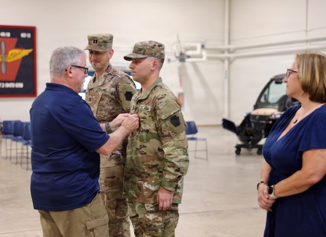A military ceremony taking place inside what appears to be a National Guard or Army aviation hangar facility. A civilian man in a blue polo shirt is pinning something onto the uniform of a soldier in Army Combat Uniform (ACU/OCP pattern), while a second uniformed soldier with captain's rank insignia stands nearby. A woman in a navy dress observes the ceremony from the right side.
