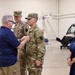 A military ceremony taking place inside what appears to be a National Guard or Army aviation hangar facility. A civilian man in a blue polo shirt is pinning something onto the uniform of a soldier in Army Combat Uniform (ACU/OCP pattern), while a second uniformed soldier with captain's rank insignia stands nearby. A woman in a navy dress observes the ceremony from the right side.