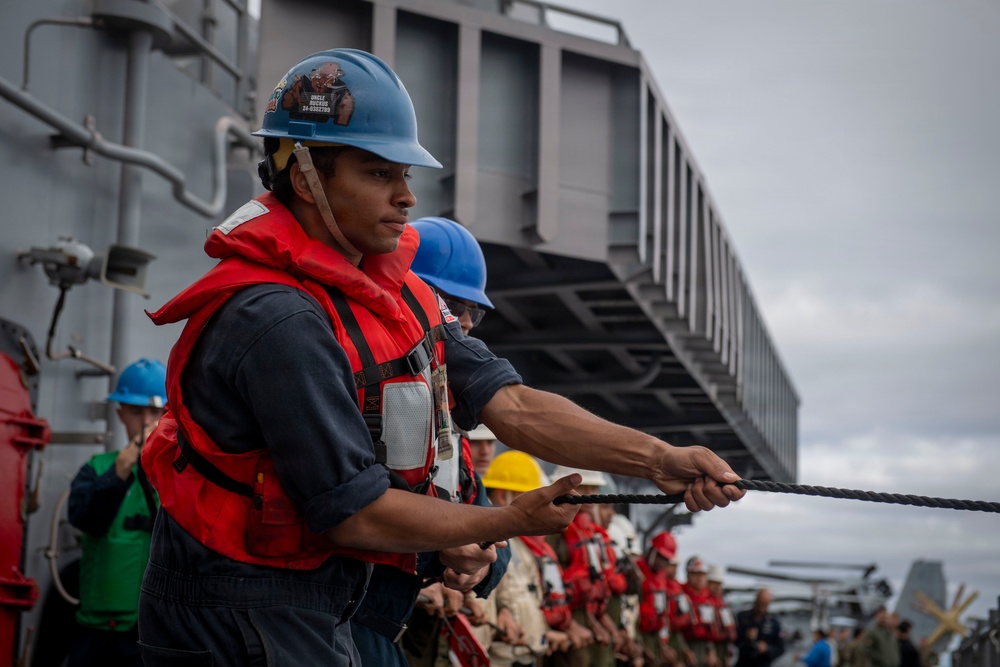 A group of naval personnel are engaged in a line-handling evolution aboard a military vessel, pulling a thick mooring or replenishment rope in unison. The foreground sailor wears a blue hard hat with stickers, a red life vest over a navy blue uniform, and is gripping a dark twisted rope with both hands. Multiple crew members in various colored hard hats and red life vests are lined up behind, suggesting a coordinated seamanship drill or underway replenishment operation.