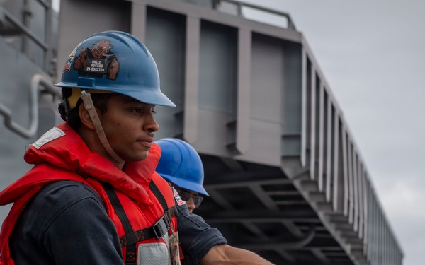 A group of naval personnel are engaged in a line-handling evolution aboard a military vessel, pulling a thick mooring or replenishment rope in unison. The foreground sailor wears a blue hard hat with stickers, a red life vest over a navy blue uniform, and is gripping a dark twisted rope with both hands. Multiple crew members in various colored hard hats and red life vests are lined up behind, suggesting a coordinated seamanship drill or underway replenishment operation.