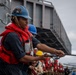 A group of naval personnel are engaged in a line-handling evolution aboard a military vessel, pulling a thick mooring or replenishment rope in unison. The foreground sailor wears a blue hard hat with stickers, a red life vest over a navy blue uniform, and is gripping a dark twisted rope with both hands. Multiple crew members in various colored hard hats and red life vests are lined up behind, suggesting a coordinated seamanship drill or underway replenishment operation.