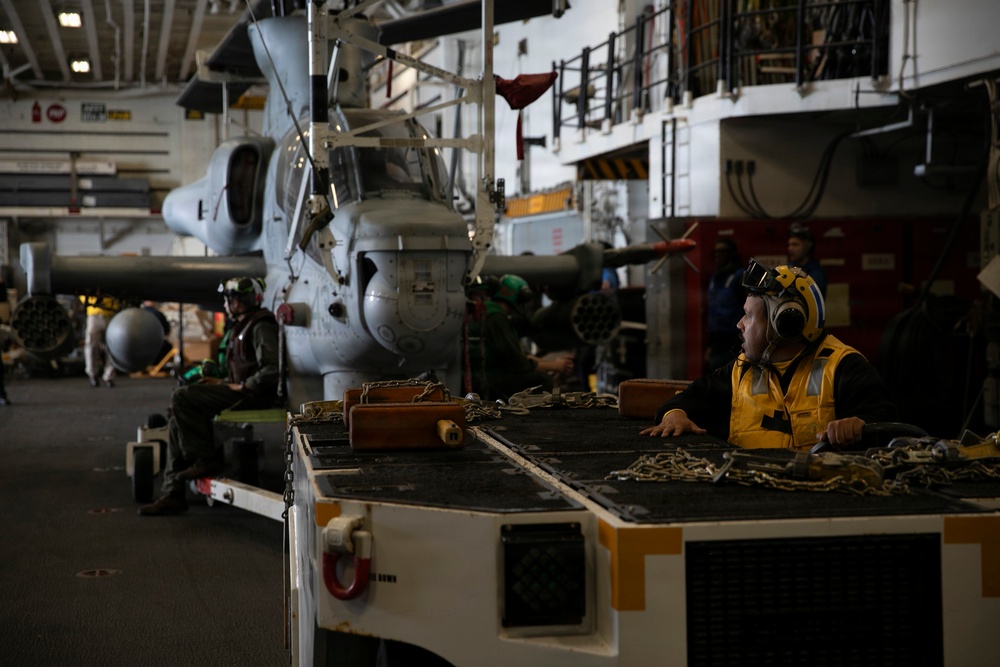 The image shows the interior hangar bay of a naval vessel, likely an aircraft carrier or amphibious assault ship, with military helicopters stored and personnel working. A crew member in a yellow safety vest and hearing protection is visible in the foreground near a equipment cart loaded with chains and tie-down gear. The environment is industrial and dimly lit, consistent with a ship's hangar deck during maintenance or aircraft handling operations.