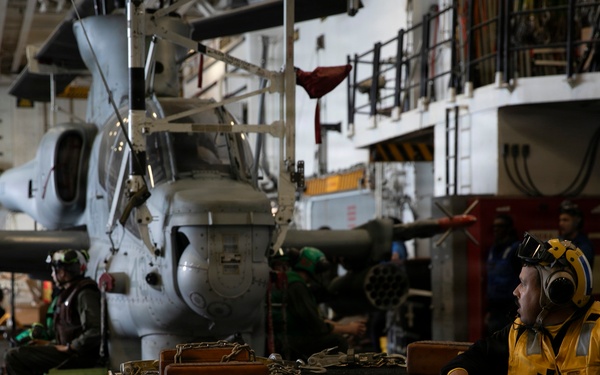 The image shows the interior hangar bay of a naval vessel, likely an aircraft carrier or amphibious assault ship, with military helicopters stored and personnel working. A crew member in a yellow safety vest and hearing protection is visible in the foreground near a equipment cart loaded with chains and tie-down gear. The environment is industrial and dimly lit, consistent with a ship's hangar deck during maintenance or aircraft handling operations.
