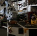 The image shows the interior hangar bay of a naval vessel, likely an aircraft carrier or amphibious assault ship, with military helicopters stored and personnel working. A crew member in a yellow safety vest and hearing protection is visible in the foreground near a equipment cart loaded with chains and tie-down gear. The environment is industrial and dimly lit, consistent with a ship's hangar deck during maintenance or aircraft handling operations.