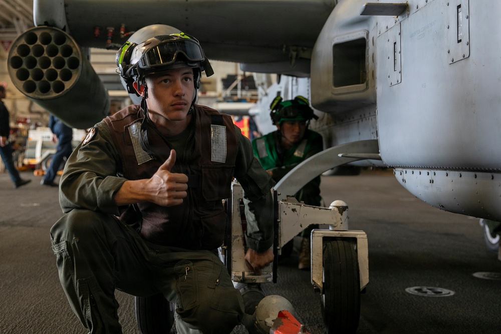 A military aviation crew member in flight gear crouches near the landing gear of an aircraft, giving a thumbs-up gesture. The setting appears to be an aircraft carrier hangar deck, with another crew member visible in the background. The aircraft has a visible rocket pod or multi-barrel weapon system mounted on it.