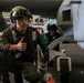 A military aviation crew member in flight gear crouches near the landing gear of an aircraft, giving a thumbs-up gesture. The setting appears to be an aircraft carrier hangar deck, with another crew member visible in the background. The aircraft has a visible rocket pod or multi-barrel weapon system mounted on it.