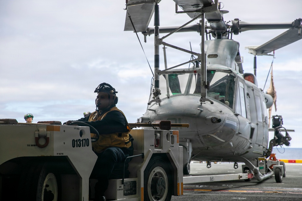 A military deck crew member operates a tow tractor (numbered 013170) to move a gray military helicopter on what appears to be a naval vessel's flight deck. The operator is wearing a helmet with ear protection and a tactical vest, consistent with flight deck crew attire. The scene takes place under overcast skies with open ocean visible in the background.