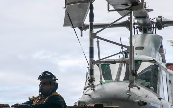 A military deck crew member operates a tow tractor (numbered 013170) to move a gray military helicopter on what appears to be a naval vessel's flight deck. The operator is wearing a helmet with ear protection and a tactical vest, consistent with flight deck crew attire. The scene takes place under overcast skies with open ocean visible in the background.