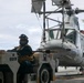 A military deck crew member operates a tow tractor (numbered 013170) to move a gray military helicopter on what appears to be a naval vessel's flight deck. The operator is wearing a helmet with ear protection and a tactical vest, consistent with flight deck crew attire. The scene takes place under overcast skies with open ocean visible in the background.
