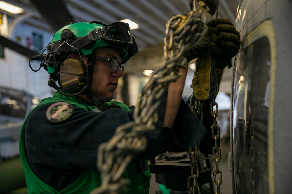 A military aviation technician wearing a green flight deck jersey and helmet is securing chains to an aircraft in what appears to be a carrier hangar bay. The individual is using tie-down chains with yellow fittings to secure the aircraft to the deck. The setting is consistent with a U.S. Navy aircraft carrier hangar deck environment.