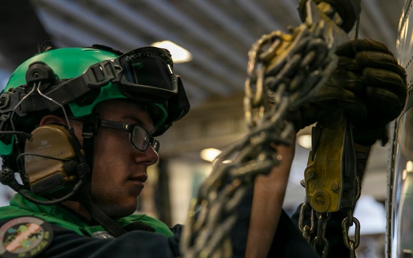 A military aviation technician wearing a green flight deck jersey and helmet is securing chains to an aircraft in what appears to be a carrier hangar bay. The individual is using tie-down chains with yellow fittings to secure the aircraft to the deck. The setting is consistent with a U.S. Navy aircraft carrier hangar deck environment.