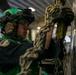 A military aviation technician wearing a green flight deck jersey and helmet is securing chains to an aircraft in what appears to be a carrier hangar bay. The individual is using tie-down chains with yellow fittings to secure the aircraft to the deck. The setting is consistent with a U.S. Navy aircraft carrier hangar deck environment.